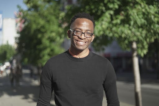 Hansel is a Black man with short dark hair and thick-rimmed glasses. He's standing outside on a street in front of tall trees, wearing a plain black top.