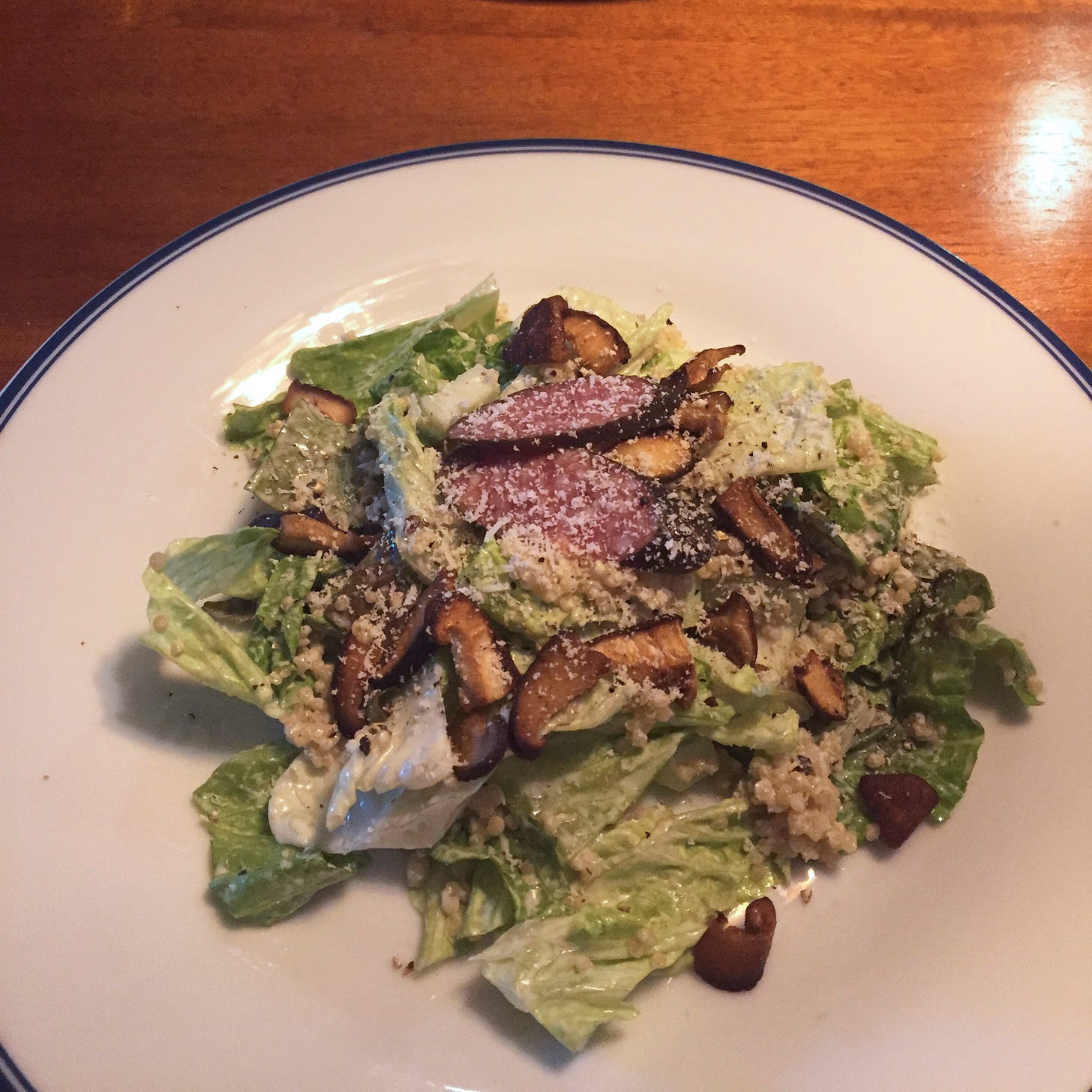 On a white plate, a quinoa Caesar salad topped with pieces of shiitake mushroom, parmesan, and two small pieces of salami.