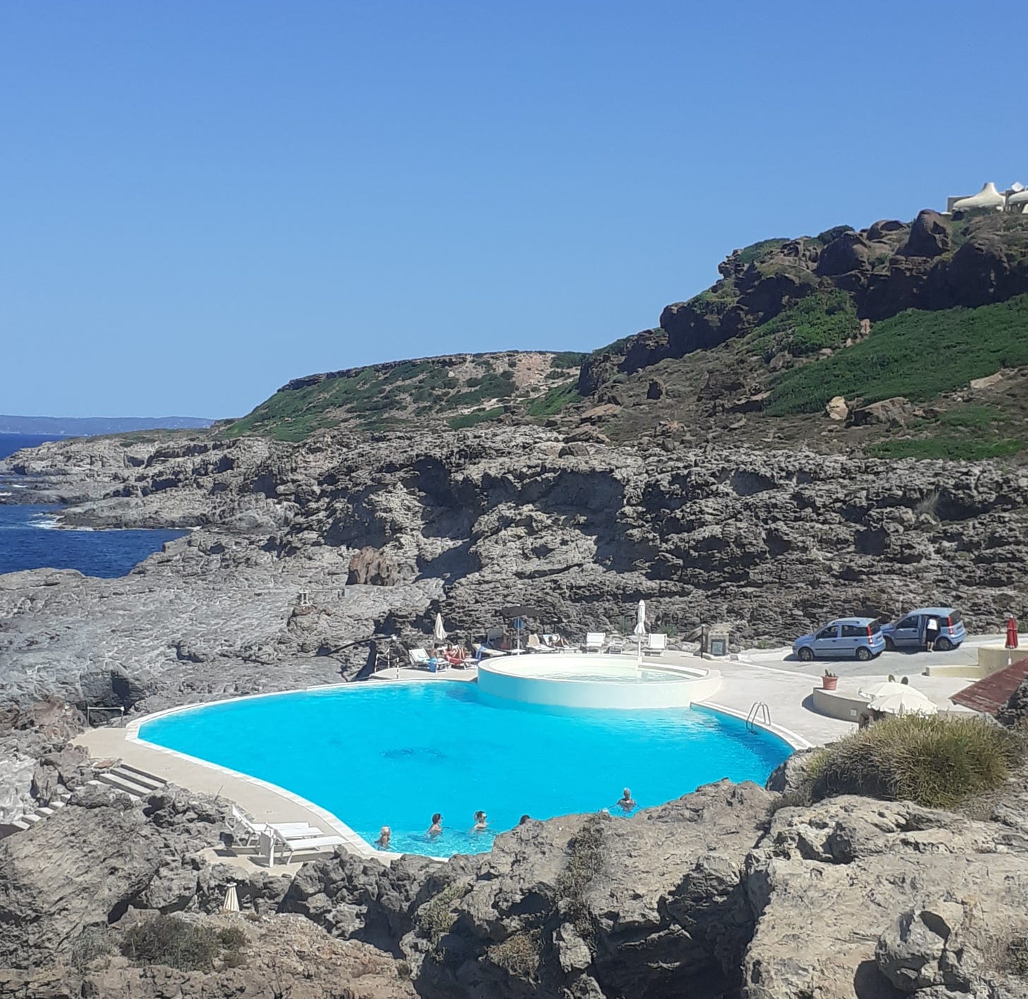 Una piscina artificiale scavata sulla roccia di fronte al mare sull'isola di Sant'Antioco: alcune persone stanno facendo il bagno mentre altre prendono il sole nelle sedie a sdraio a bordo piscina.