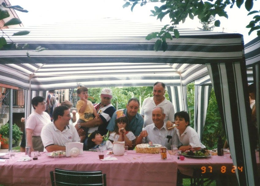 An Italian-Canadian family posing by a cake under a white and green striped tent in the backyard. The garden is visible behind them.