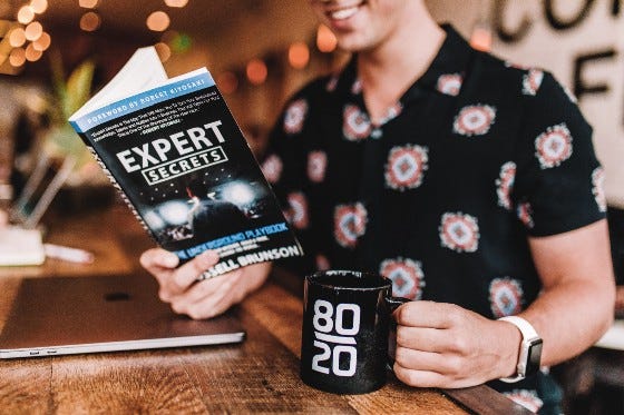 person drinking cofee reading a book titled “expert secrets”