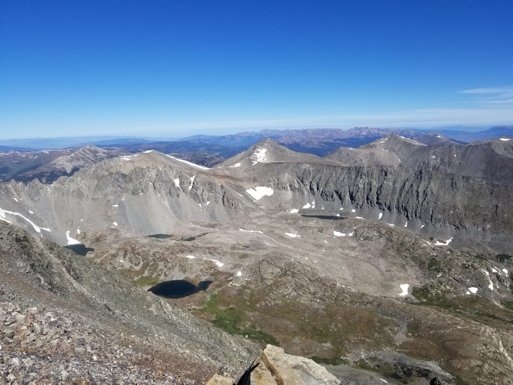 The view from Quandary's summit. A glacial basin ringed by craggy, gray mountains sits beneath a clear blue sky. Several snowmelt lakes dot the bottom. From one of these lakes, a small river runs back down off the mountain.