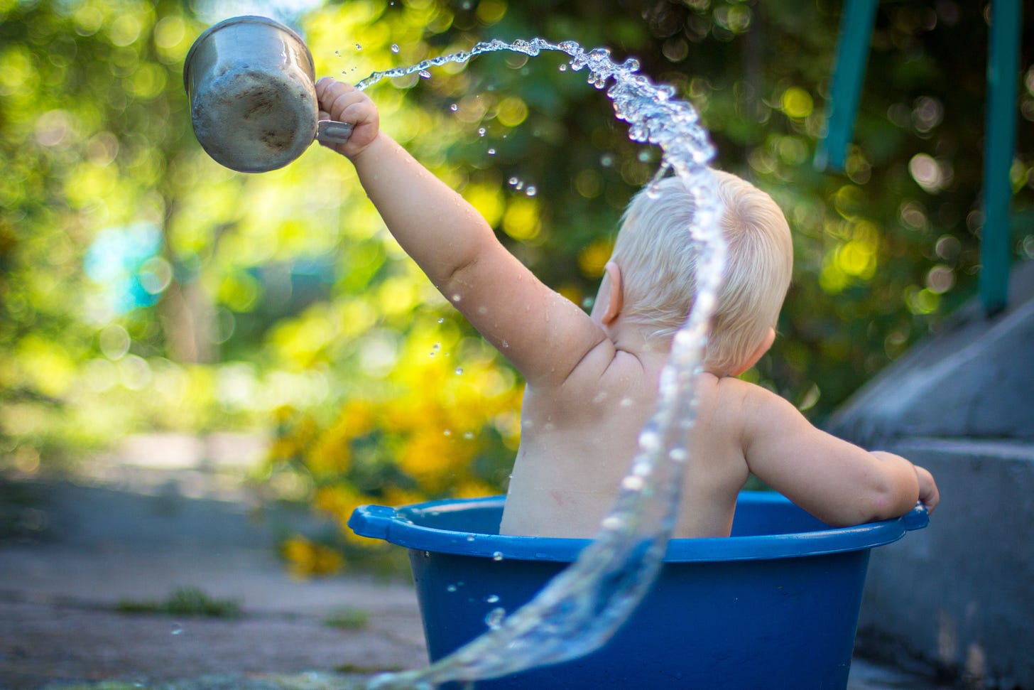 A toddler sat in a bowl of water splashes himself with water from a tin cup. Photo by Lubomirkin on Unsplash
