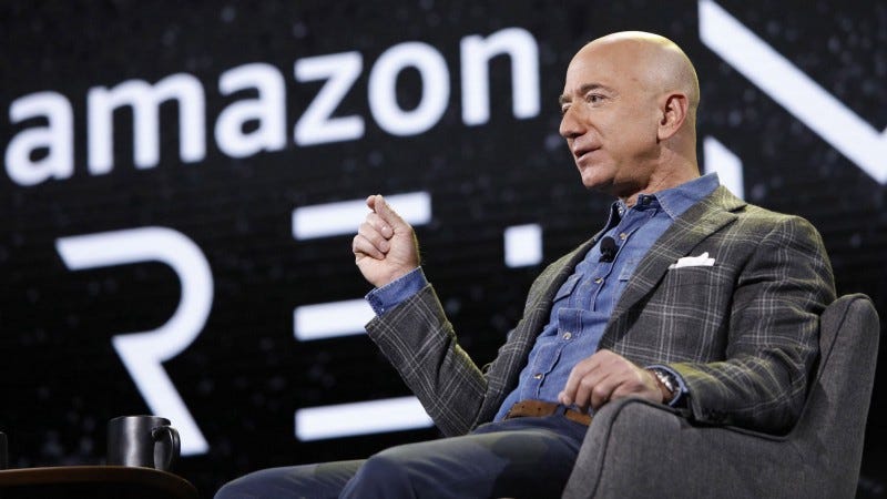 Jeff Bezos seated in a chair with the Amazon logo behind him.