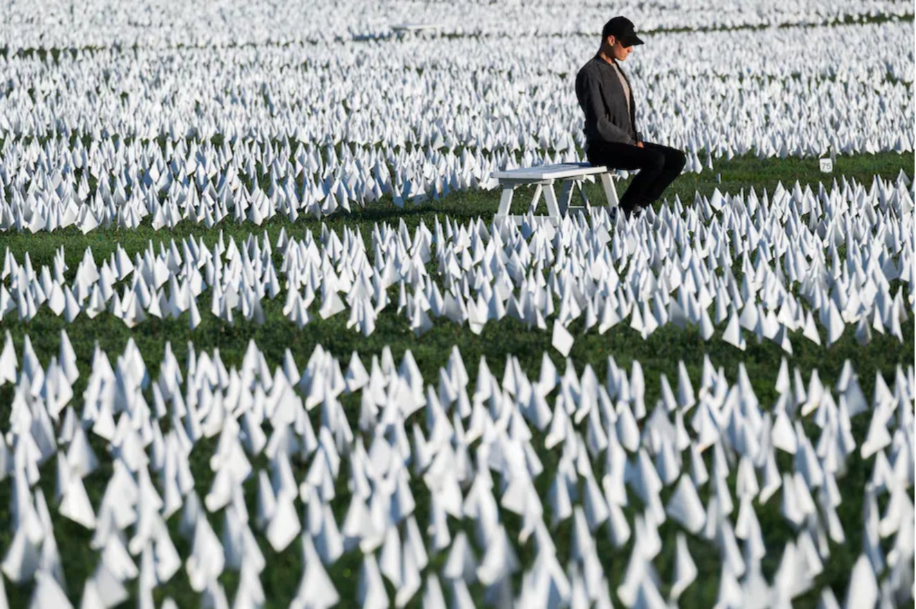 A sea of small white flags as far as the eye can see. A man in a baseball hat sits on a bench in the middle of the installation.