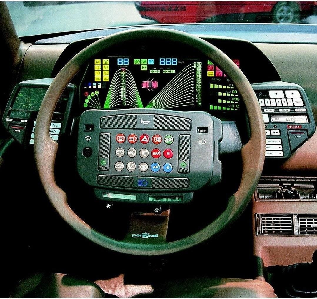 Imagem mostra painel de um carro, com volante repleto de botões, além de painel com mais botões e um visor com informações gráficas coloridas.
