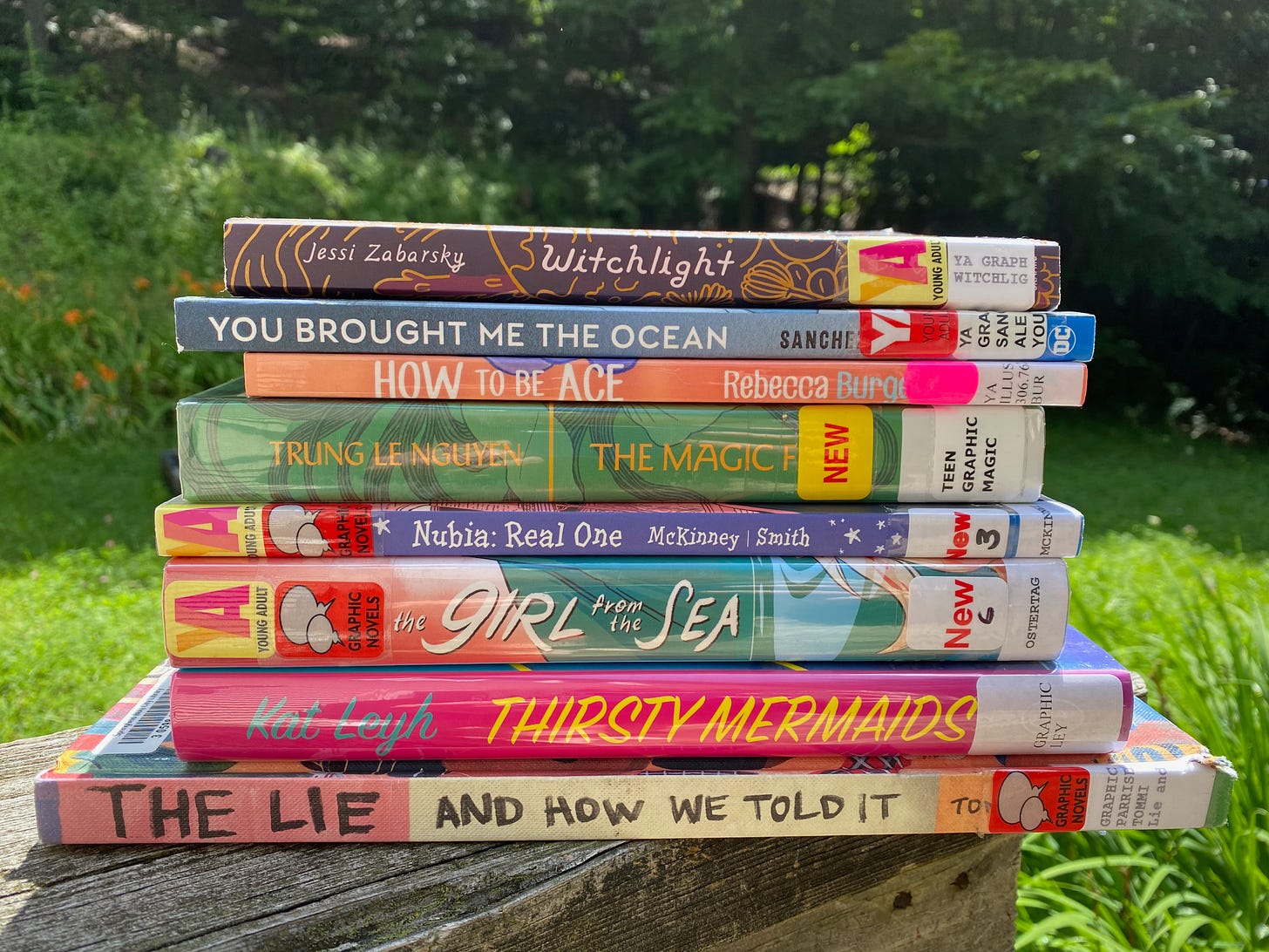 A stack of graphic novels sitting on a wooden railing in front of a sunny lawn: The Lie and How We Told It, Thirsty Mermaids, The Girl from the Sea, Nubia, The Magic Fish, How to be Ace, You Brought Me the Ocean, and Witchlight.