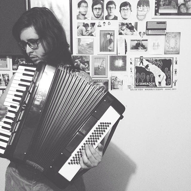 A black and white picture of Angelo playing an accordeon in front of a white wall. In the wall are hanged several pictures of him, as long as CD covers and illustrations