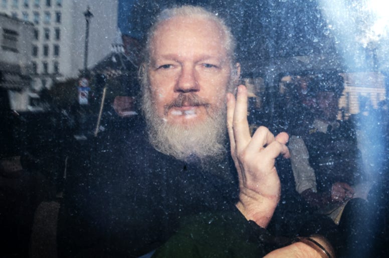 Color picture of Julian Assange from behind the window of a car. He is a white male, appearing to be somewhere in his 50s, wearing black clothes, has white hair and a white beard, and is holding up two fingers in a V sign. In the reflection of the window you can see the London street with buildings, streetlight, and street signs.