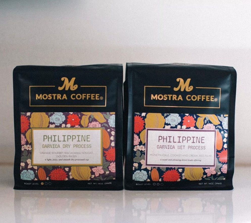 Two bags of Mostra Coffee with their name and logo in gold on black. A flower design is in a box with text indicating the coffee inside is from the Philippines.