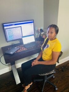 Candace Milburn sits in front of a computer monitor and keyboard, looking at the camera. She wears a yellow t-shirt and black pants.