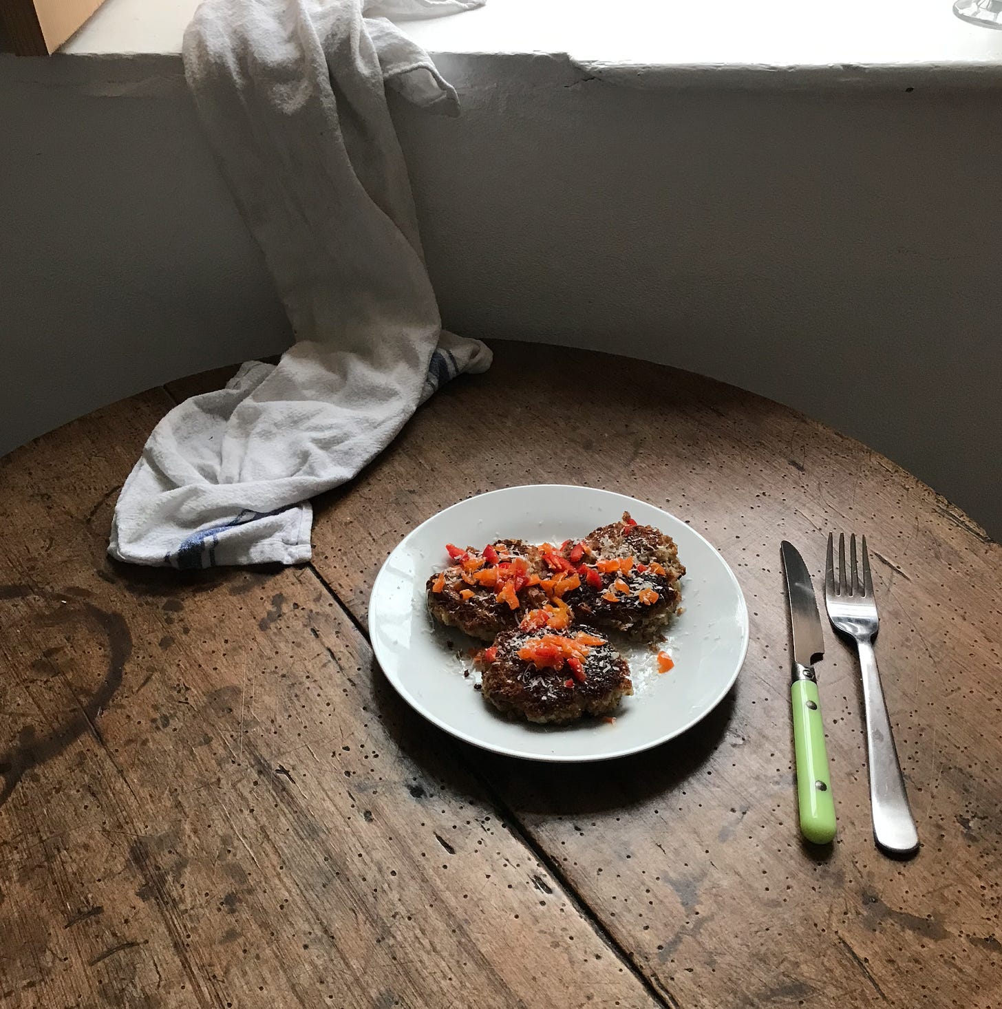 A white plate with three rough rounds, topped with something red, peppers cubed, and white gratings of parmesan cheese. To the side of the plate is a metal knife and fork, the knife has a bright green plastic handle. The plate is on an antique wooden table, and a tea towel half sits, half drapes over the table, in the background. 