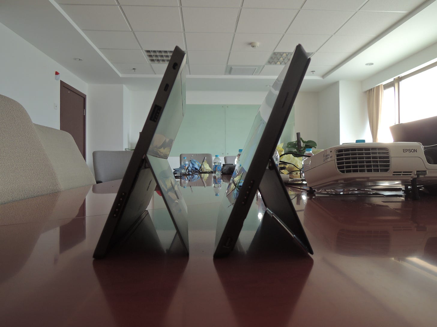Surface and Surface Pro, or GT and GTX. Both are positioned on a conference room table with the kickstands out. It can be seen that GTX is much thicker than GT.