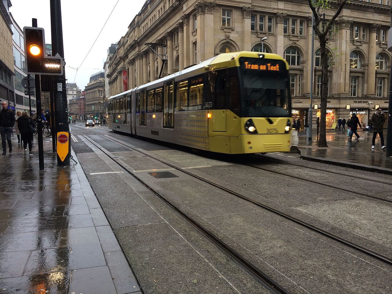 A test tram runs along the Manchester Metrolink Second City Crossing on the street. Its a wet day, with a few people walking to and from shops in the background.