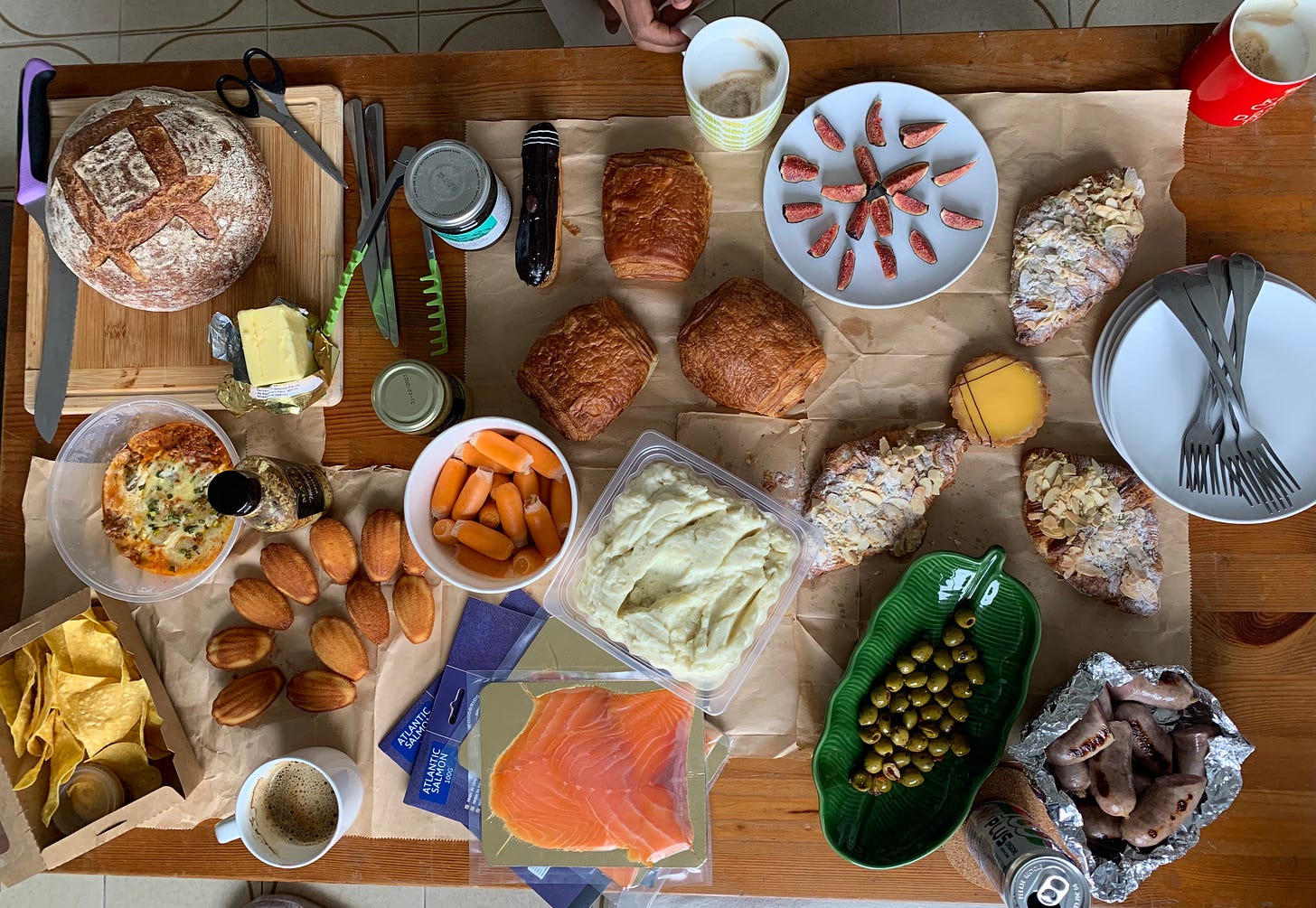 A brown wooden table with an assortment of food ranging from a round sourdough loaf, mashed potatoes, sausages, olives, almost croissants, figs, a lemon tart, three chocolat au pain, madeleines, nachos and dip, and smoked salmon.