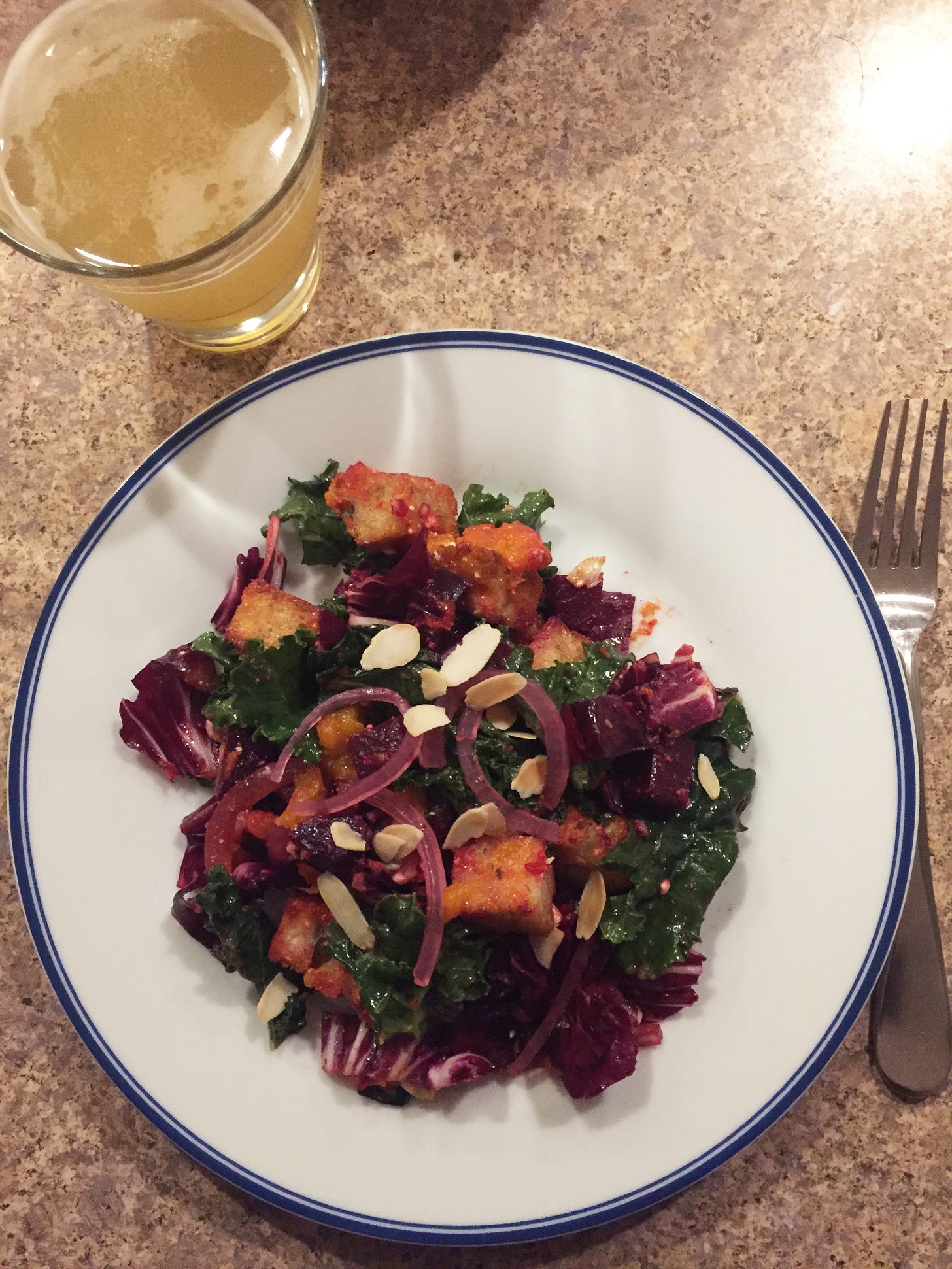 A plate of panzanella with beets and pieces of roasted squash among croutons, kale, and radicchio. On top are toasted sliced almonds and pickled red onion. A glass of beer sits to the upper left corner of the plate.