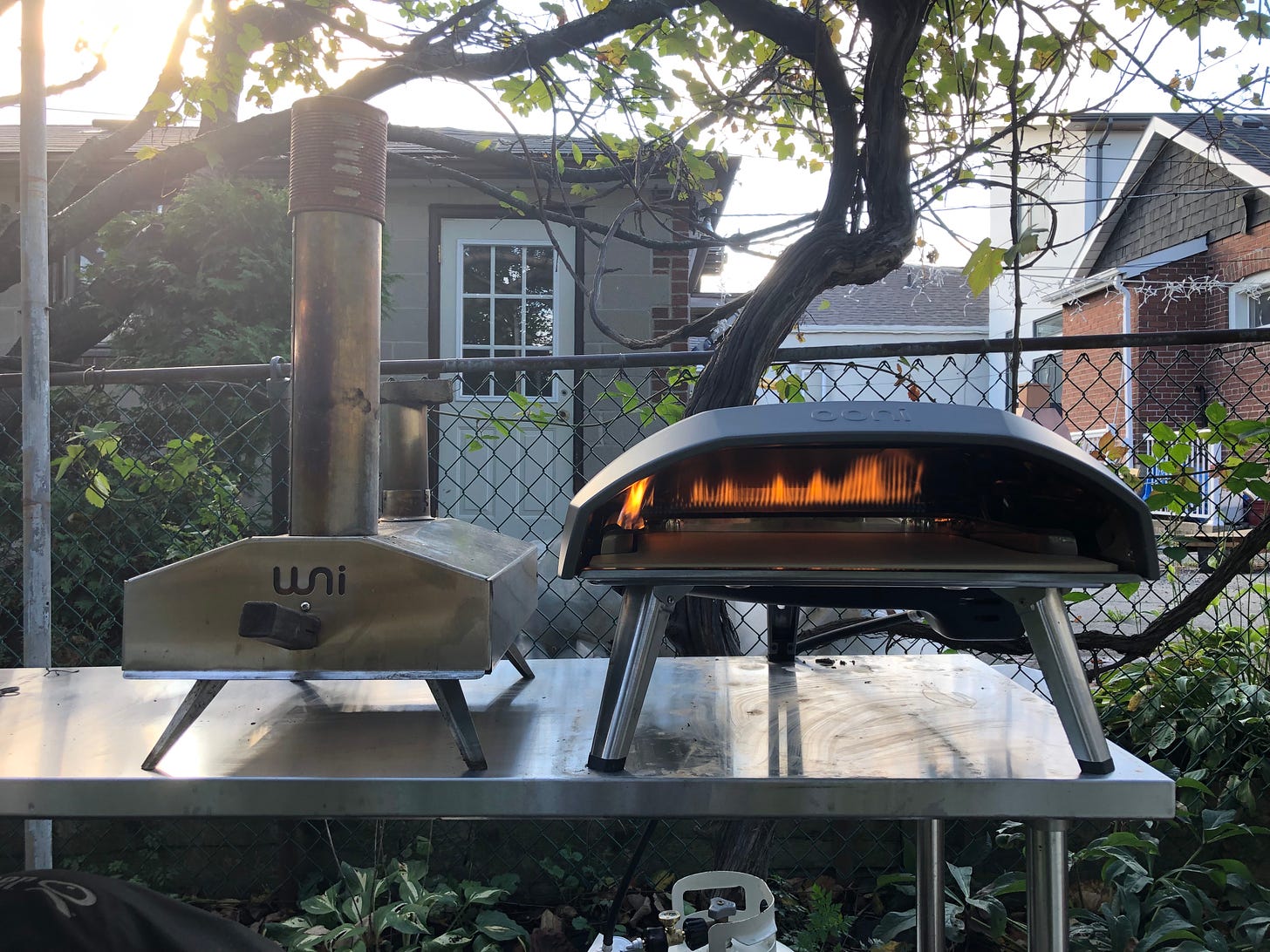 A small, wood pellet-fired Uuni 2 pizza oven beside a larger, gas-fired Ooni Koda 16 on a steel table. 