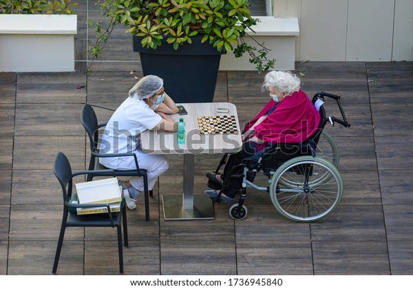 PARIS, FRANCE - MAY 20, 2020: nurse playing checkers with an elderly and disabled woman in a nursing home during the coronavirus pandemic Covid-19. Both wear a protective face mask.