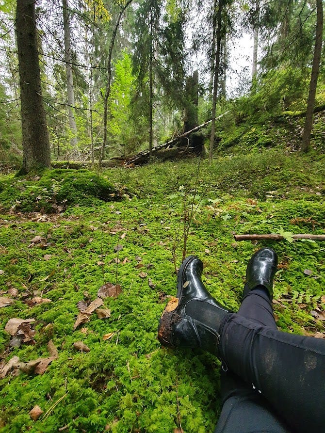 um par de pernas e botas pretas sujas de terra, cruzadas, no chão da floresta. árvores ao fundo