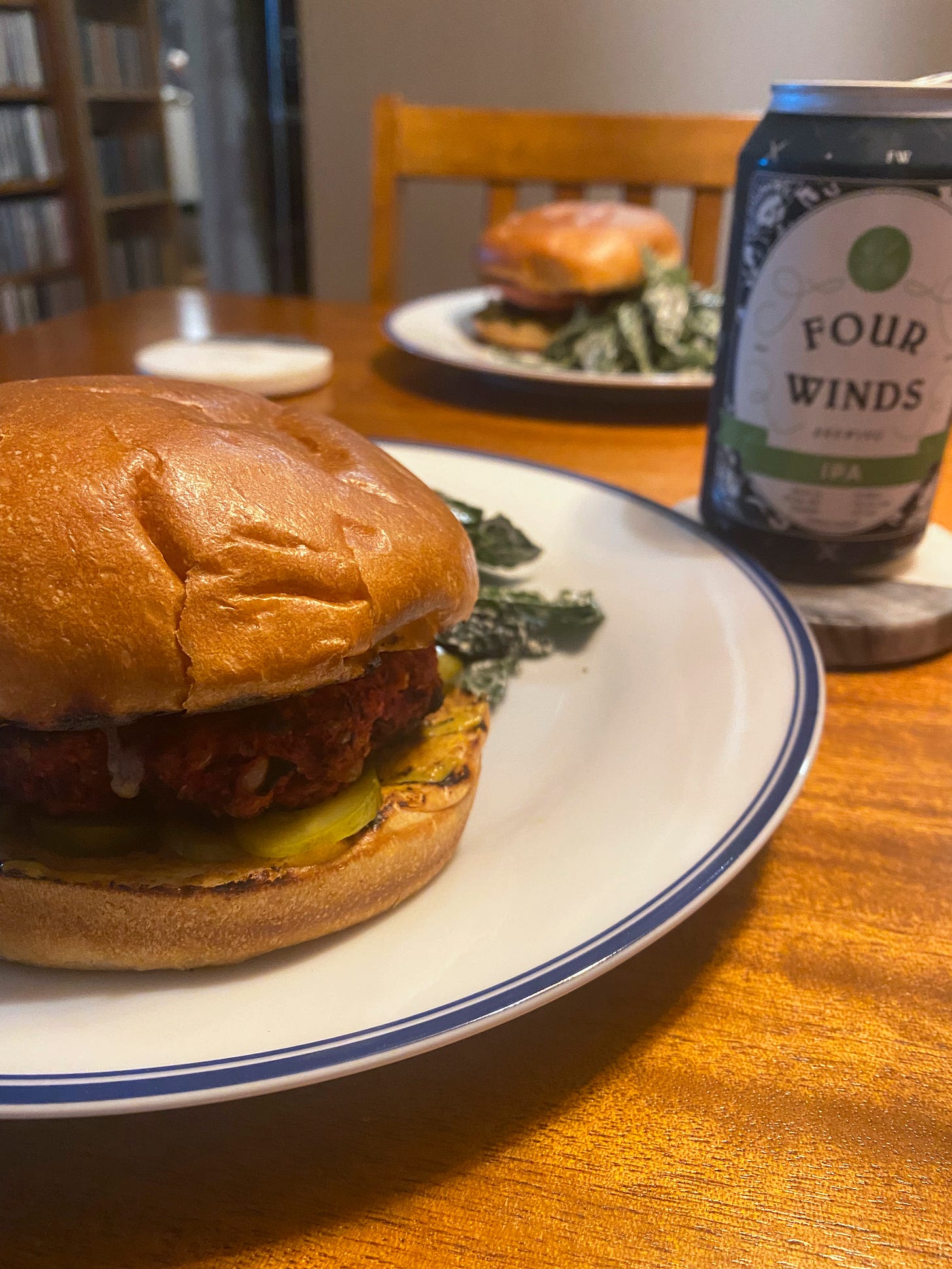 A veggie burger on a brioche bun sits on a white plate. Another plate with a burger and salad on it is in the background, and on a coaster next to the first plate is a can of Four Winds IPA.