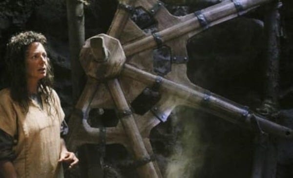 Mother (Allison Janney) stands beside an enormous wooden spoked wheel with a large spindle.