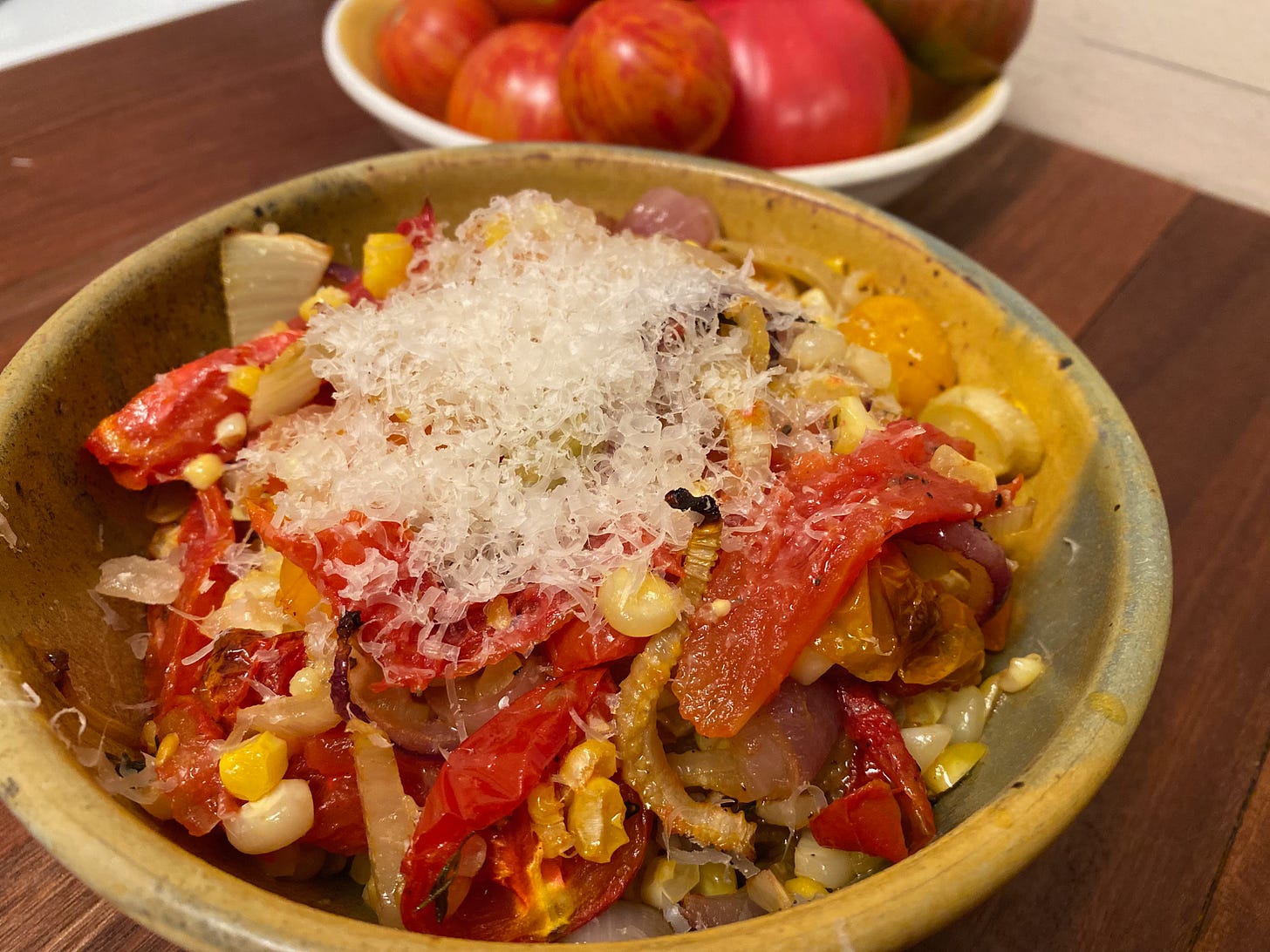 A ceramic bowl of roasted tomatoes, fennel, onions, and corn, topped with a pile of grated Parmesan cheese. The bowl sits on a wooden counter, with part of a bowl of whole tomatoes visible in the background.