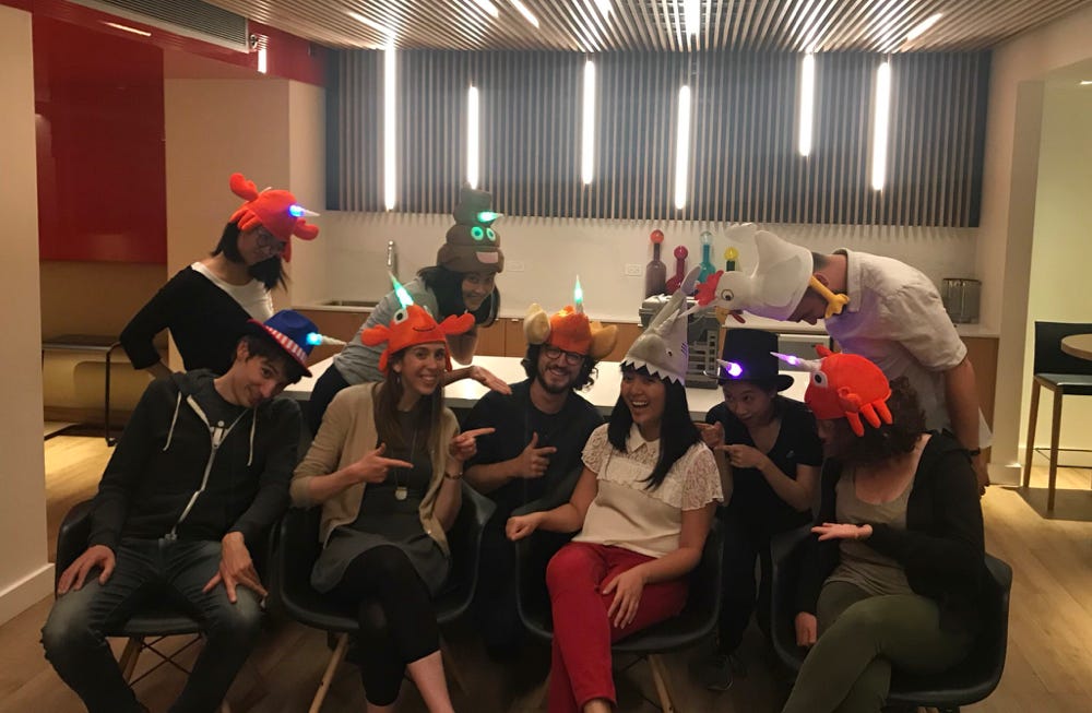 A group of 9 people with Charlyn in the center sitting down all wearing shark and emoji hats.