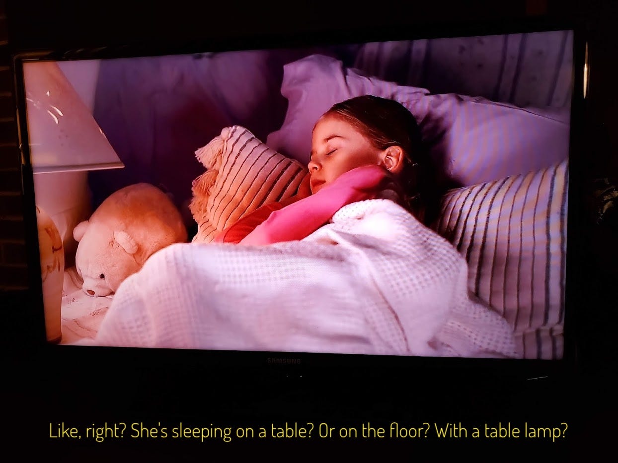 A little girl under a blanket with pillows and a stuffed animal and also a table lamp
