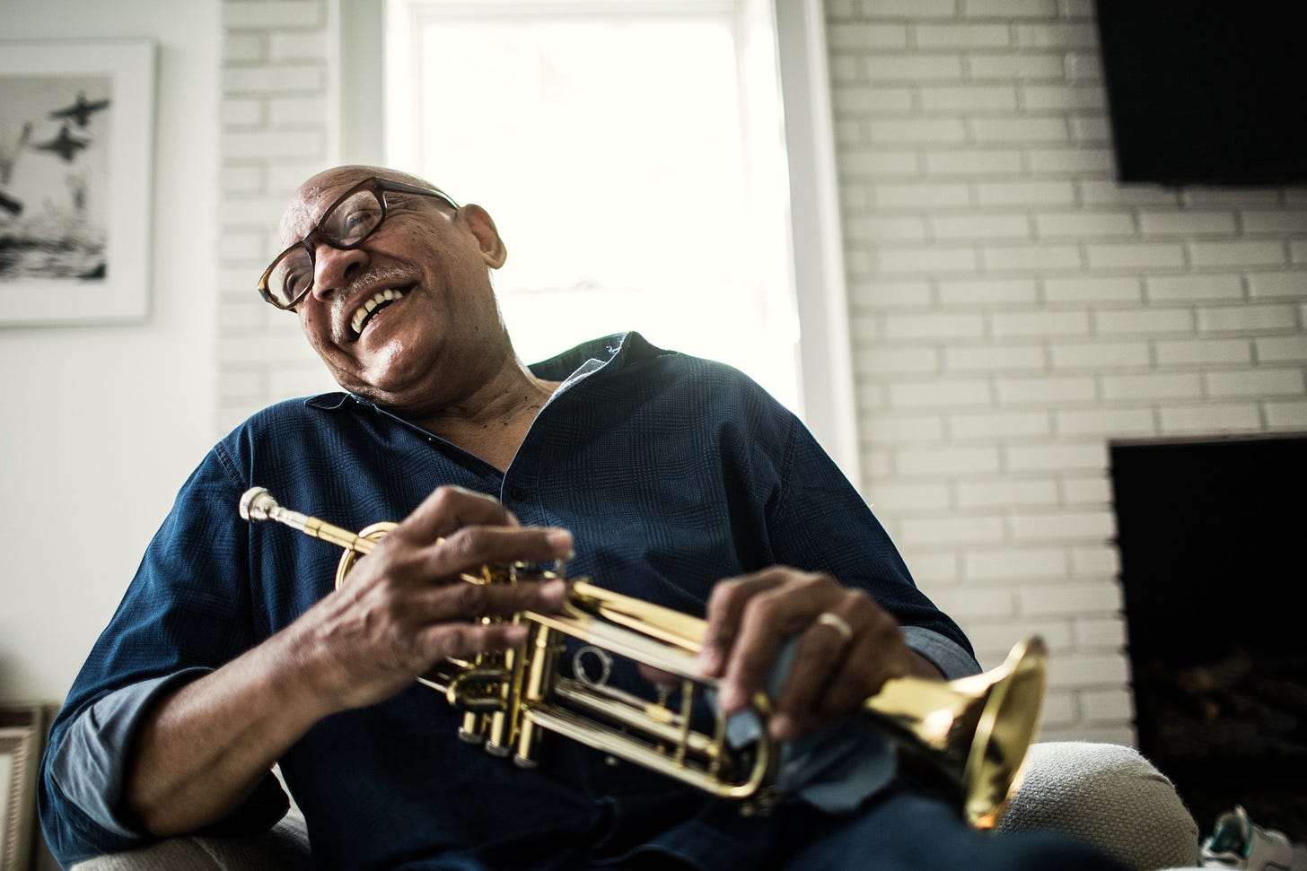 A photo of an older black man holding a trumpet in his living room. He is laughing.