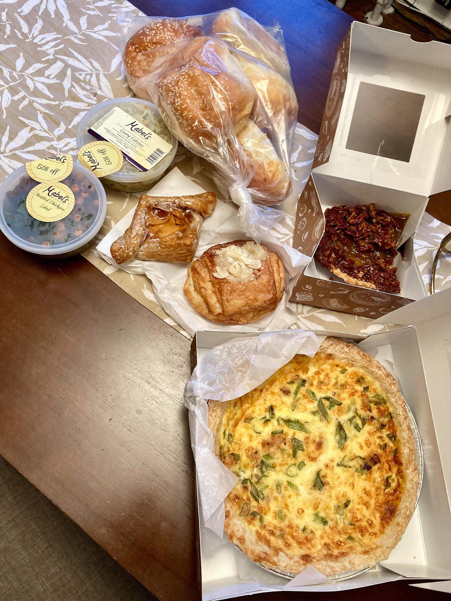 Spread of food including a full quiche, three pastries, two salads, and bag of buns