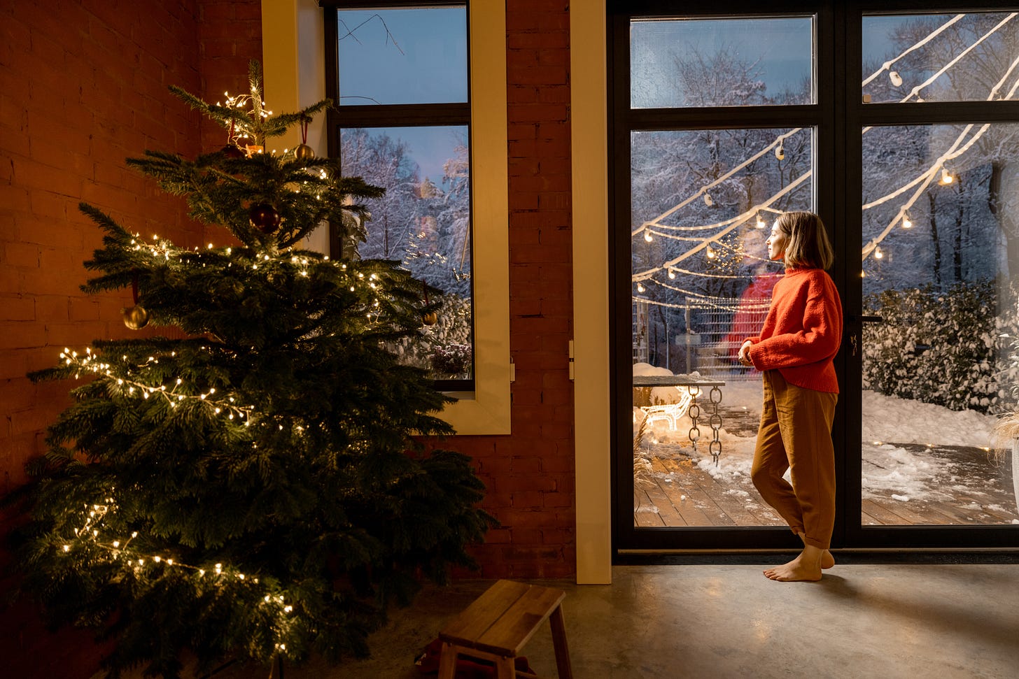 A woman stares pensively out of a cozy living room window at Christmas.