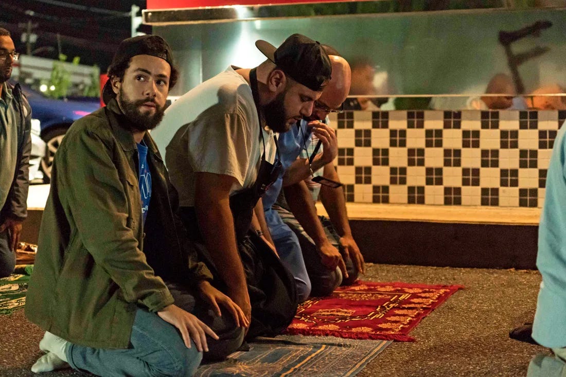 Three men kneel on prayer mats on the street at night, surrounded by other people.