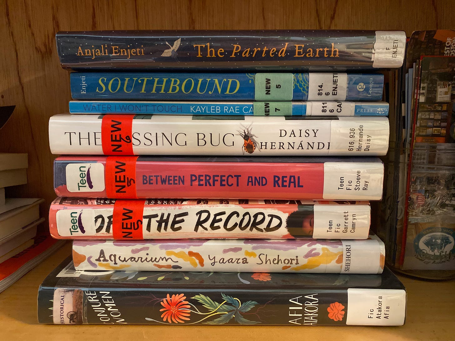 A stack of library books next to a glass jar full of bookmarks. The books are: Conjure Women, Aquarium, Off the Record, Between Perfect and Real, The Kissing Bug, Water I Won’t Touch, Southbound, and The Parted Earth.
