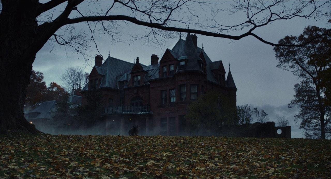 A still from the movie 'Knives Out'. A wide shot of the Thrombey mansion. The mansion is massive with turrets and made from a dark brick. The sky is grey. The trees have lost their leaves. Black dogs run through the orange leaves on the ground. The overall mood is gothic and solemn.