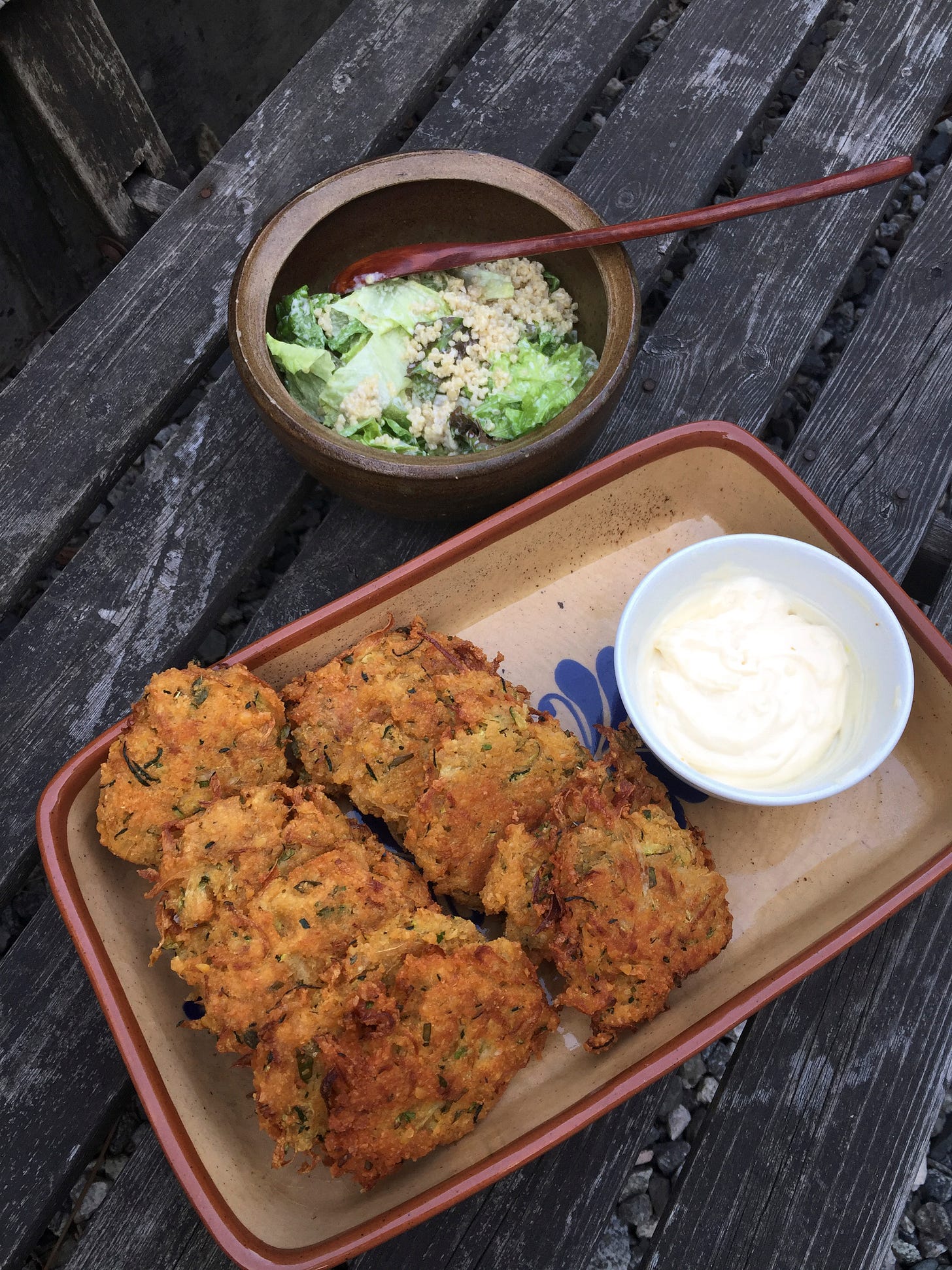 On a wooden bench, a rectangular dish full of golden-brown fritters, next to a small white dish of the lemon mayo. Behind it is a small stone bowl of quinoa Caesar salad.