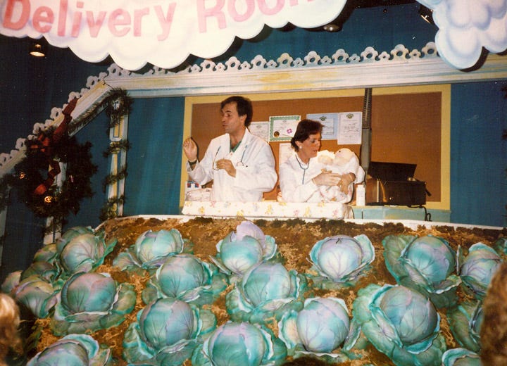 A man dressed as a doctor addresses the room as a woman dressed as a nurse coddles a Cabbage Patch Kid doll. They stand in a window behind a large fake cabbage patch. A sign reading "Delivery Room" hovers above them.