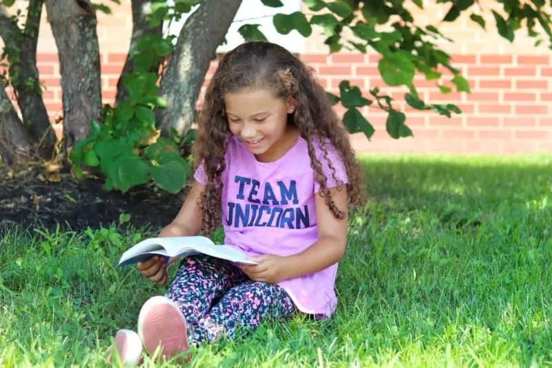 girl sitting on the grass under a tree reading a book with a team unicorn shirt on