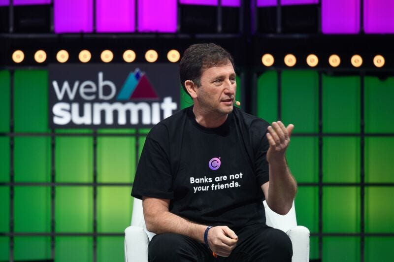 Celsius CEO Alex Mashinsky speaks at a conference while seated on a stage and wearing a T-shirt that says, 