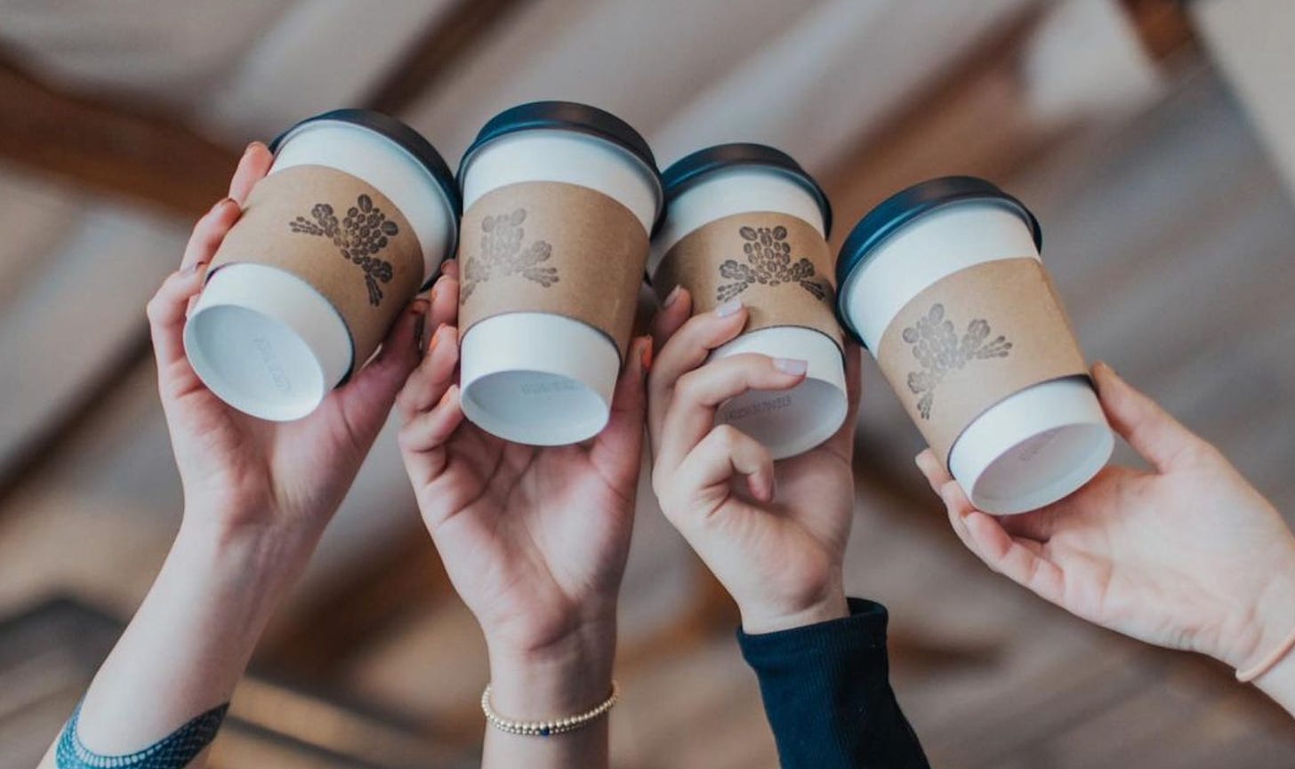 Four white hands holding up to-go coffee cups with lids. A brown coffee cup sleeve has a black Leap Coffee logo inked onto it. The cups are touching in a cheers.