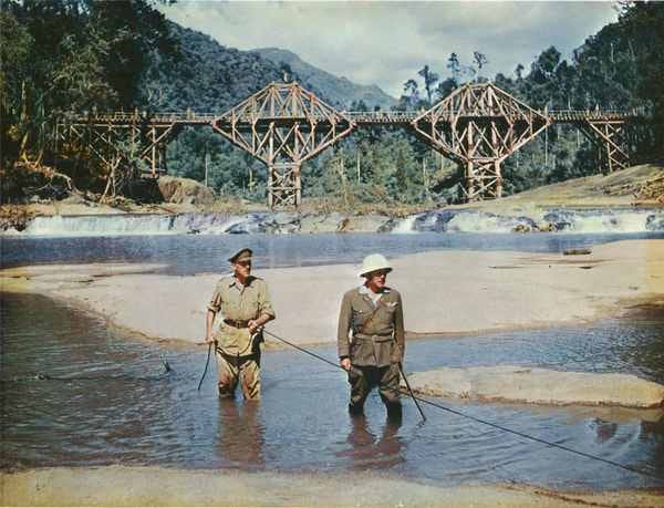 Still from the movie "The Bridge on the River Kwai" which depicts Alec Guinness following the cable powering the detonator set to blow up the titular bridge, seen in the background.