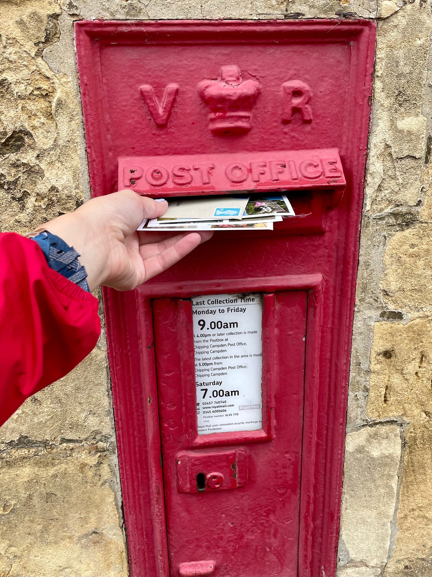 A woman puts a handful of postcards and letters into a red postbox built into a stone wall in Chipping Campden, England.