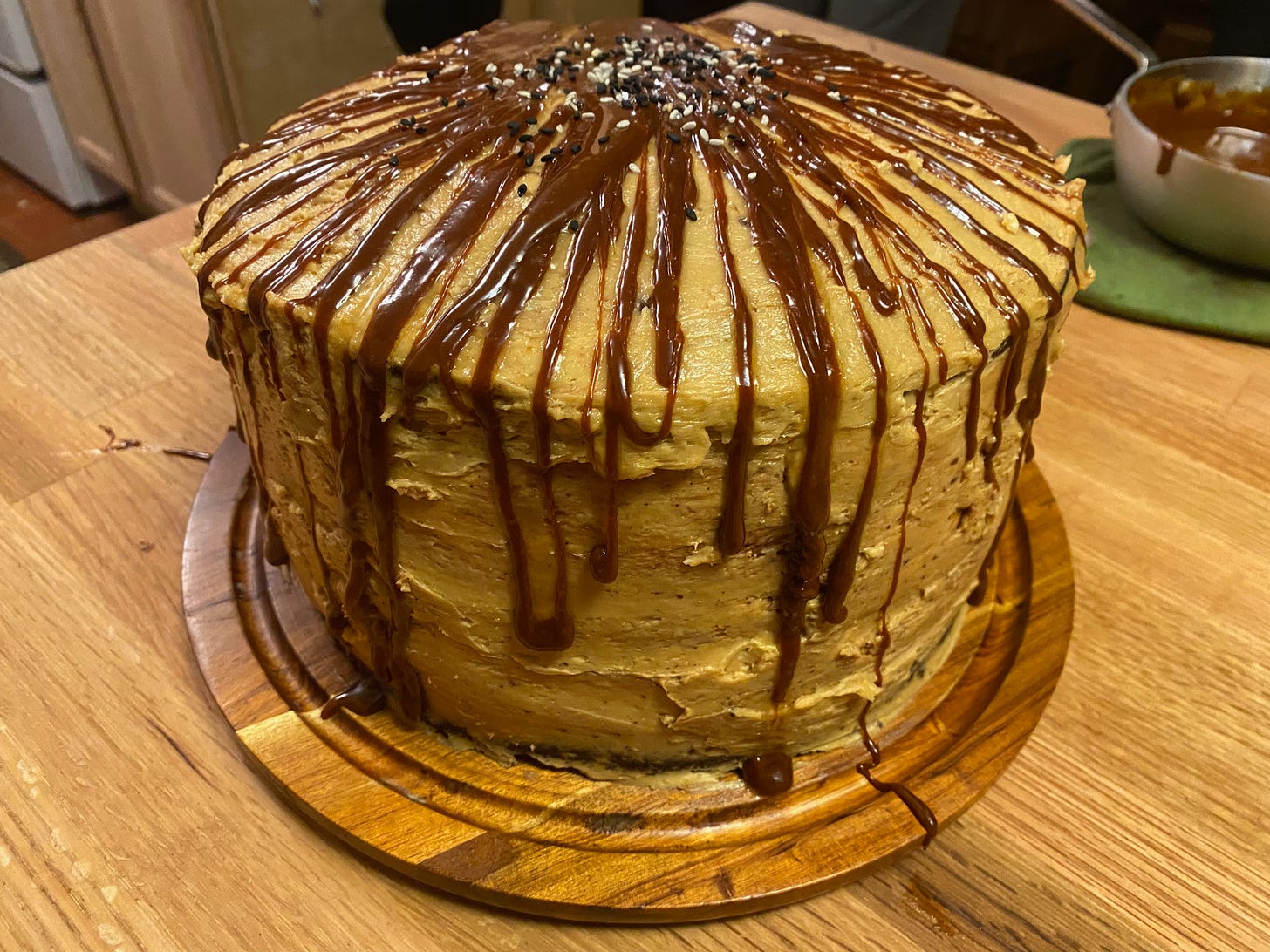 A tall layer cake sits on a kitchen counter. It’s covered in thick tahini frosting, and uneven drizzles of caramel run down the sides from the top, which is sprinkled with sesame seeds. A small pan of caramel sits on the counter behind the cake.