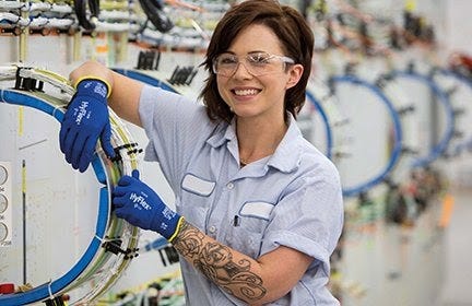 Photo of a smiling woman in a factory. She is wearing blue work gloves and protective goggles. She has a sleeve of tattoos on her arm. Seems gay.