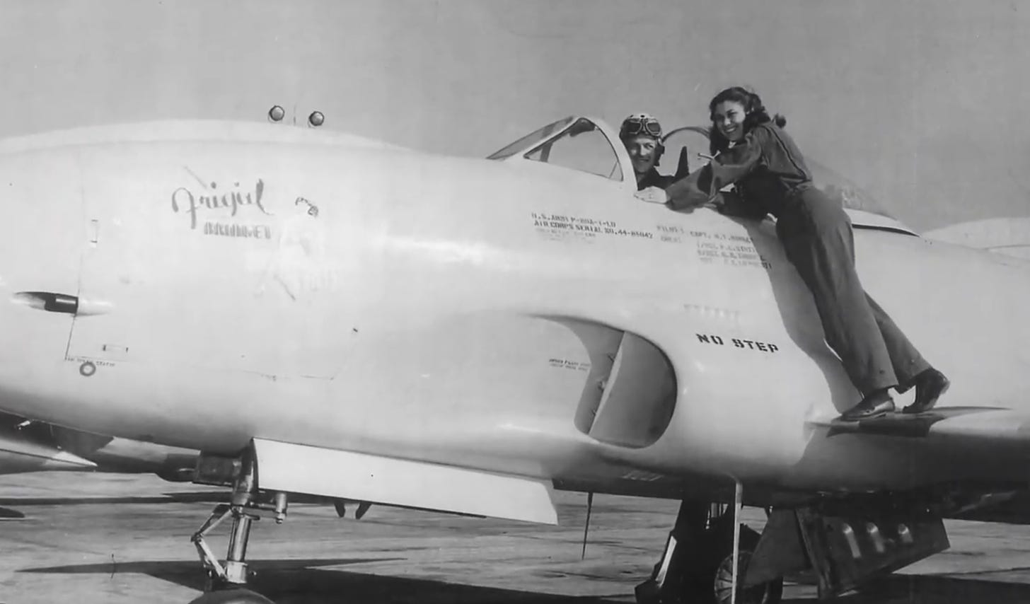 Feik stands on the wing of a plane. She is leaning towards the cockpit, smiling at the camera.  A pilot is also posing for the camera.