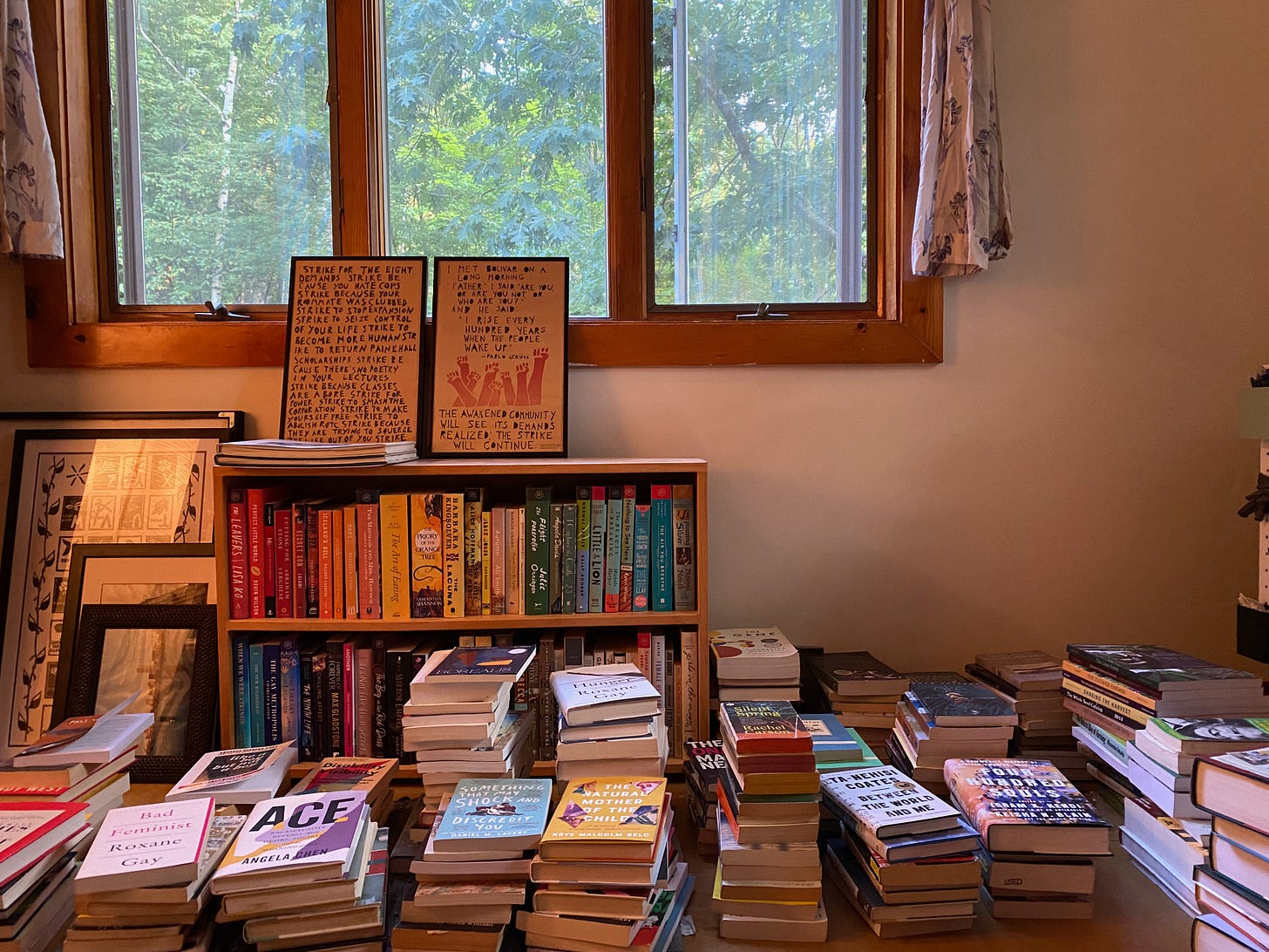 A floor covered with stacks and stacks of books. There is a bookcase against the wall under a window, filled with books arranged in a rainbow pattern, and several pieces of art lean next to it and on top of it.