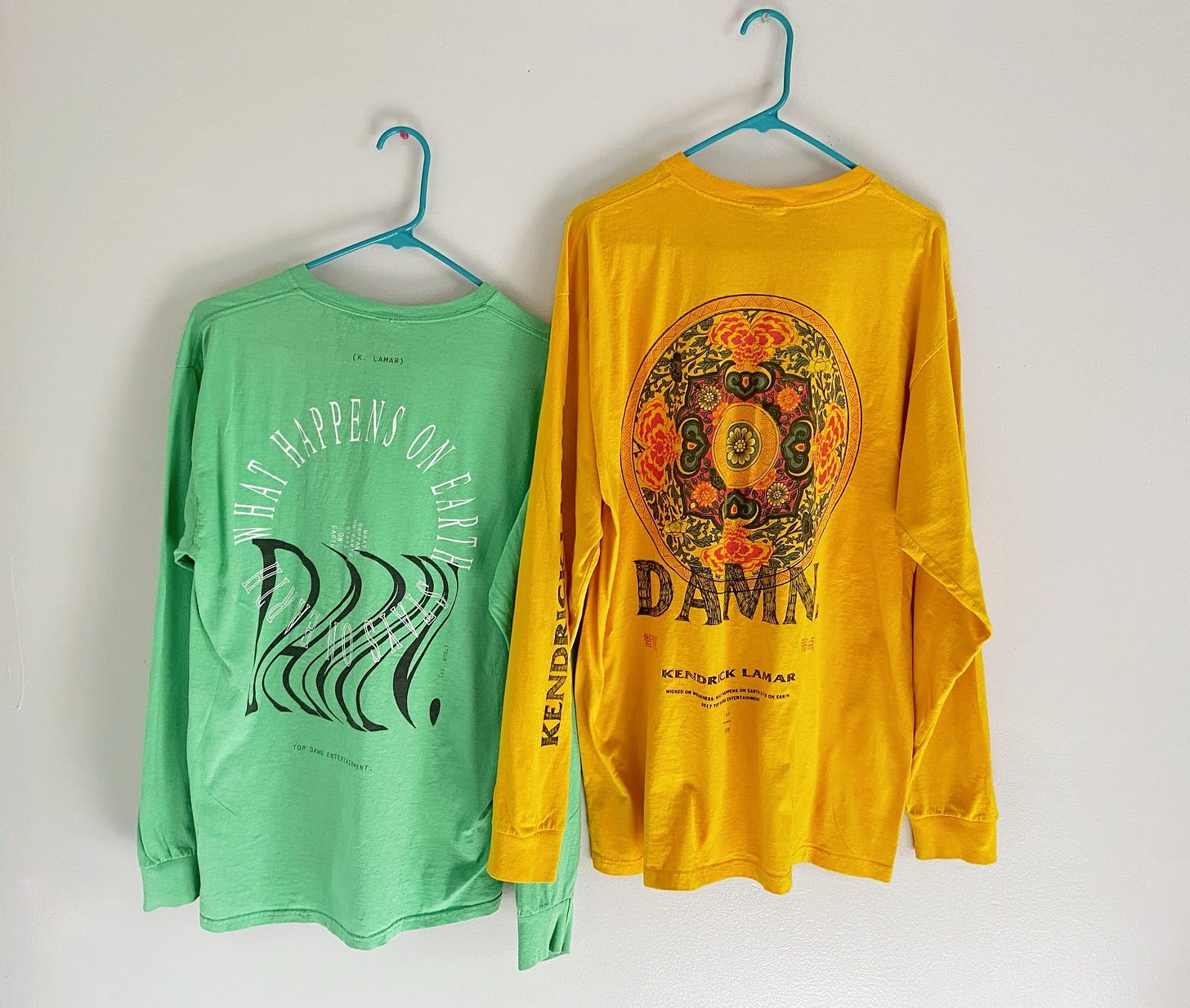 Two shirts from Kendrick Lamar's DAMN. tour, one seafoam green, the other mustard yellow. they have circle motifs and overlapping words to say "DAMN" and "What happens on Earth stays on Earth."