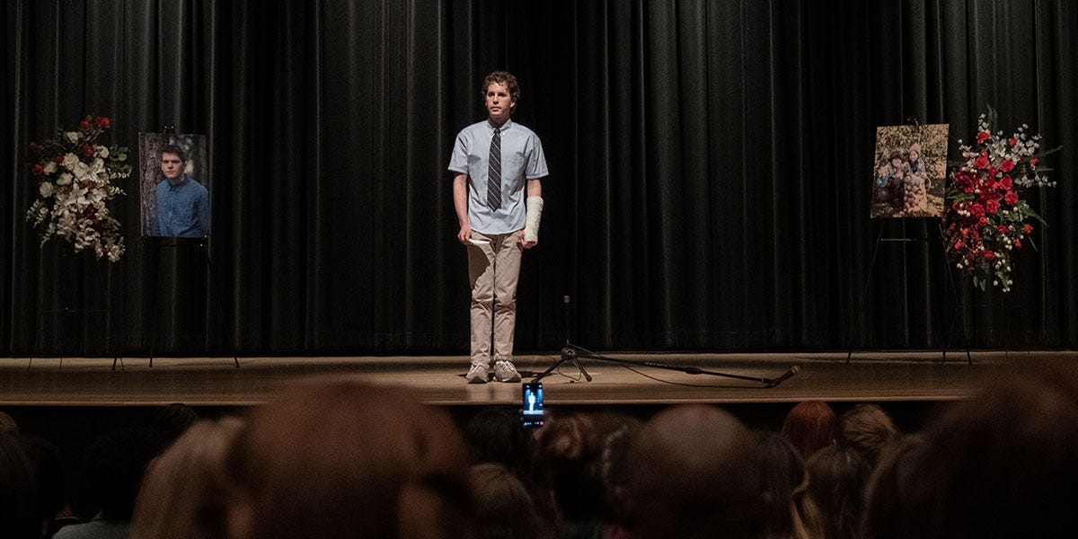 Ben Platt as Evan Hansen stands alone on an auditorium stage at a memorial for a fellow student, as other students film him with their cellphones.