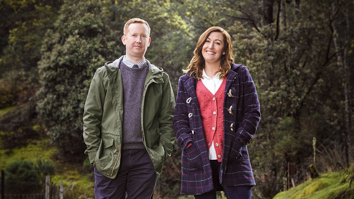 Luke McGregor and Celia Pacquola stand wearing coats with their hands in their pockets surrounded by lush green forest and grass.