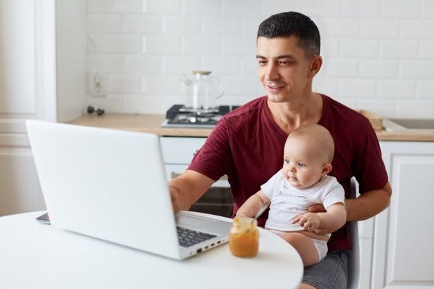 Pleasant looking man wearing maroon casual t shirt, young adult father sitting at table in kitchen in front of laptop computer looking at notebook display with positive expression. Free Photo