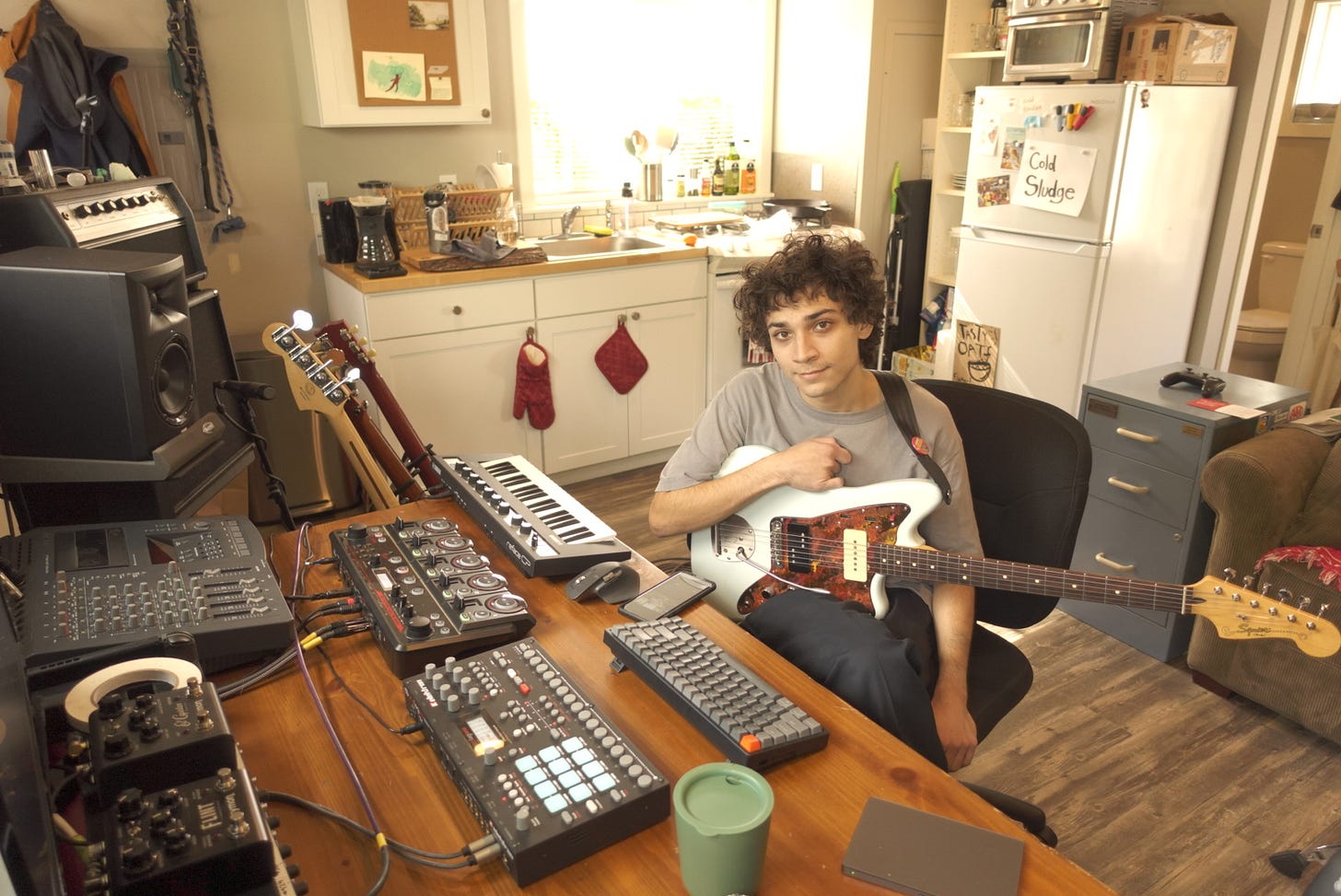 A man holding a guitar sitting in a kitchen surrounded by instruments and recording gear